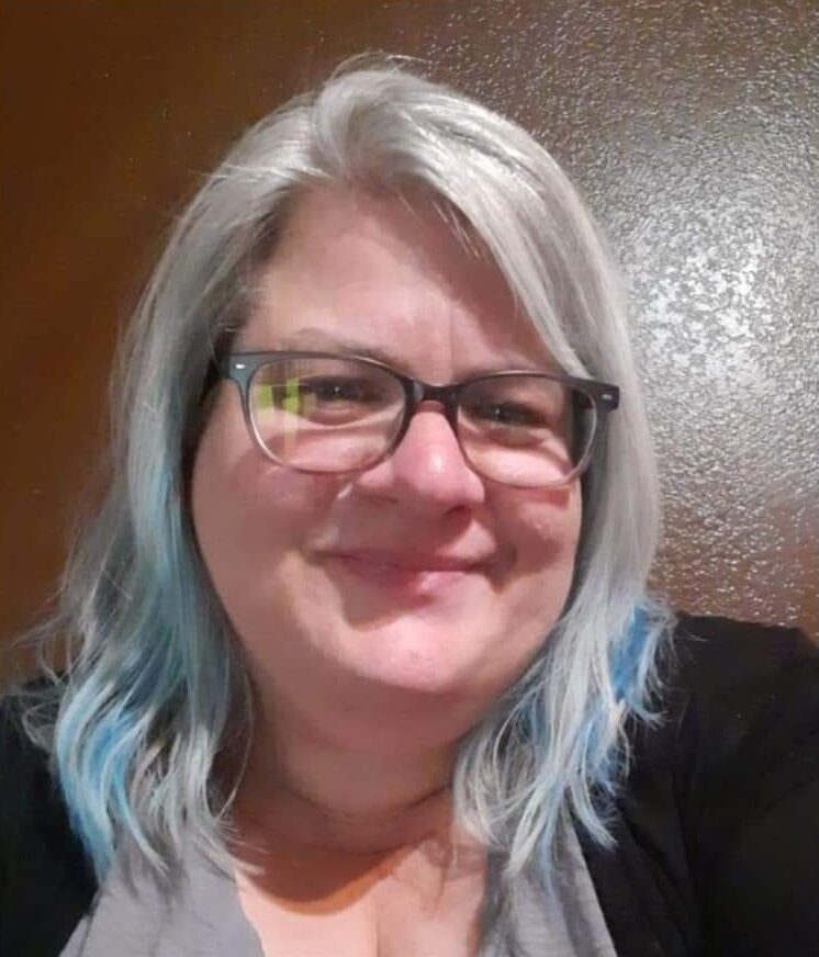 A headshot of a smiling woman with gray hair and glasses against a brown backdrop