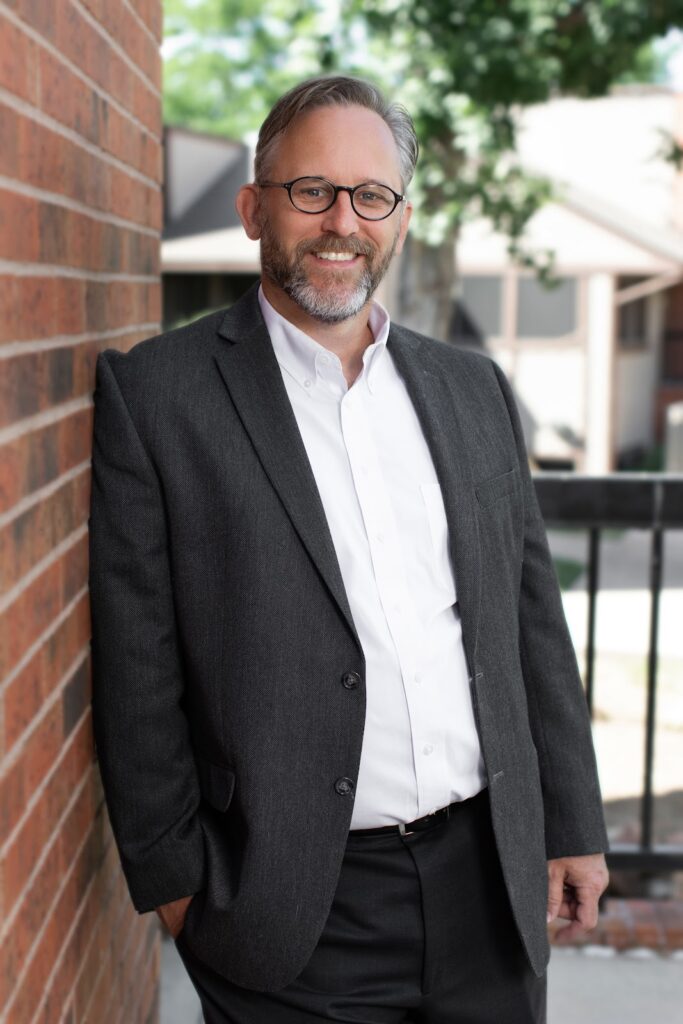 A smiling man with a salt-and-pepper beard wearing a suit, leaning against a brick wall
