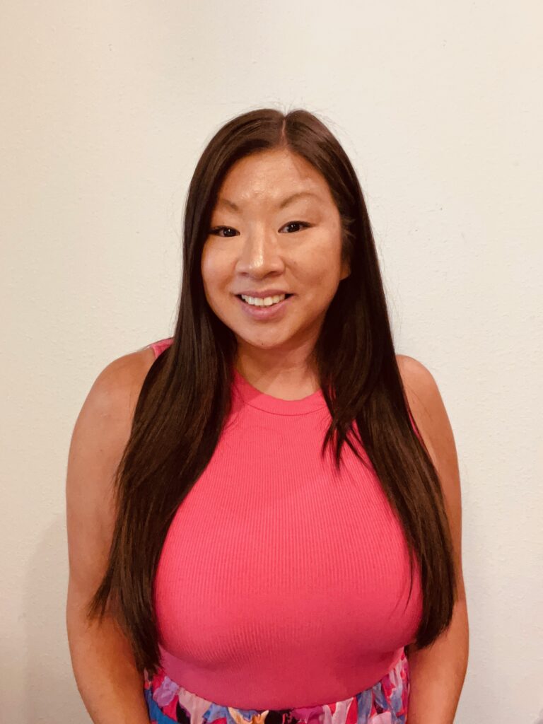 A smiling woman wearing a pink shirt against a tan backdrop