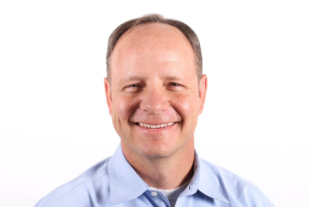 A smiling balding man wearing a dress shirt against a white backdrop